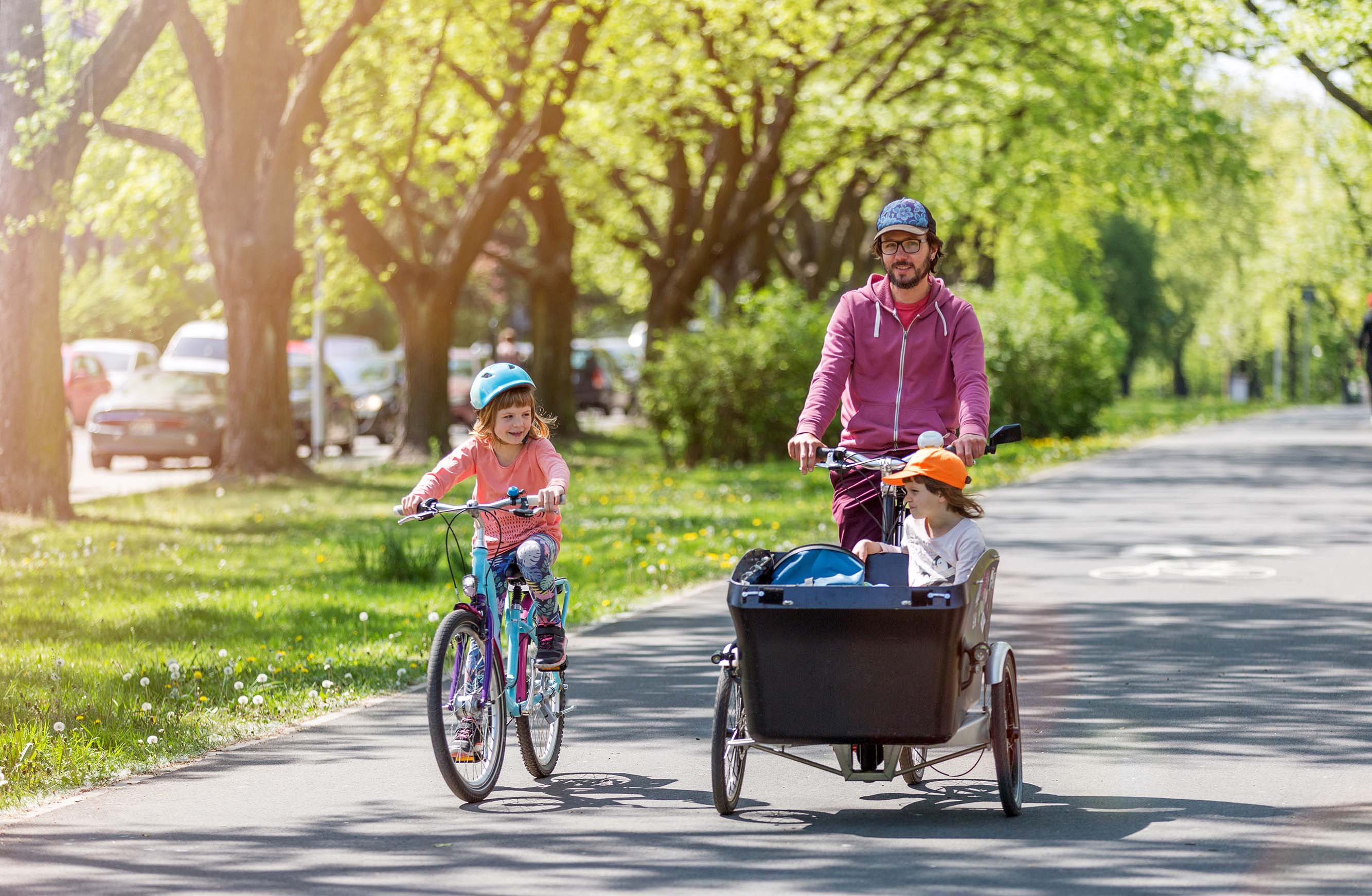 Ein Vater fährt mit einem Lastenrad auf einem Radweg, ein Kind fährt neben ihm auf einem Fahrrad. Ein zweites Kind sitzt im Lastenrad. Es ist sonnig, im Hintergrund sind geparkte Autos und Bäume zu sehen.