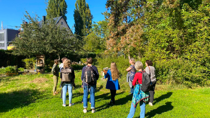 Eine Gruppe Menschen steht mit dem Rücken zur Kamera in einem Garten an einem herbstlichen, sonnigen Tag.