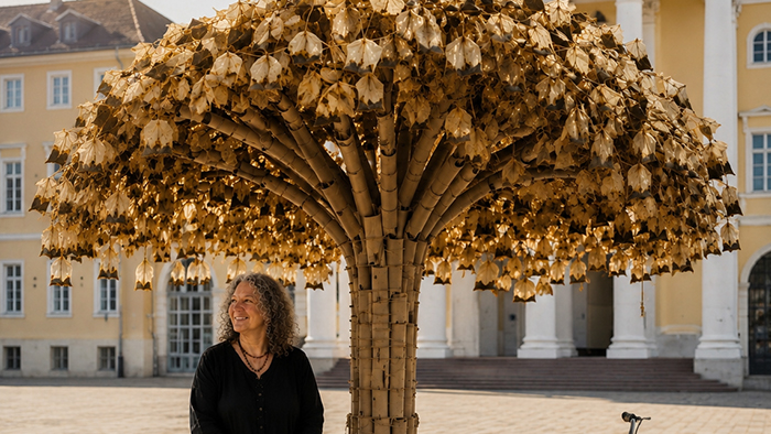 Die Künstlerin Helene Renate Schweizer sitzt neben ihrem Kunstwerk aus alten Teebeuteln, das aussieht wie ein großer Baum.